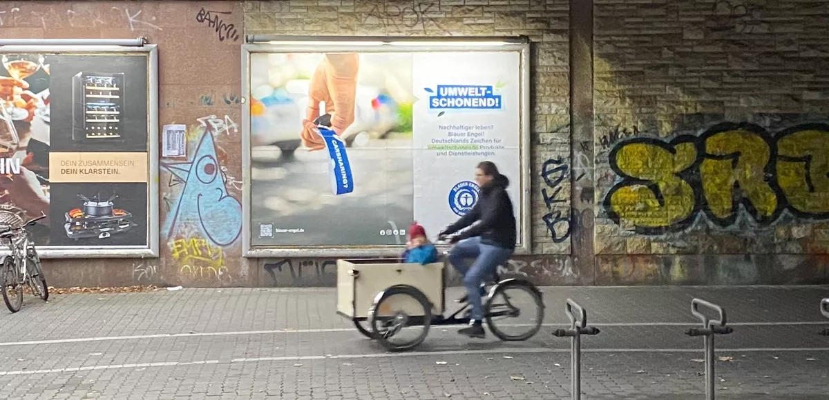 Mann fährt ein Kind in einem Lastenfahrrad an einem Werbeplakat mit dem Blauen Engel vorbei