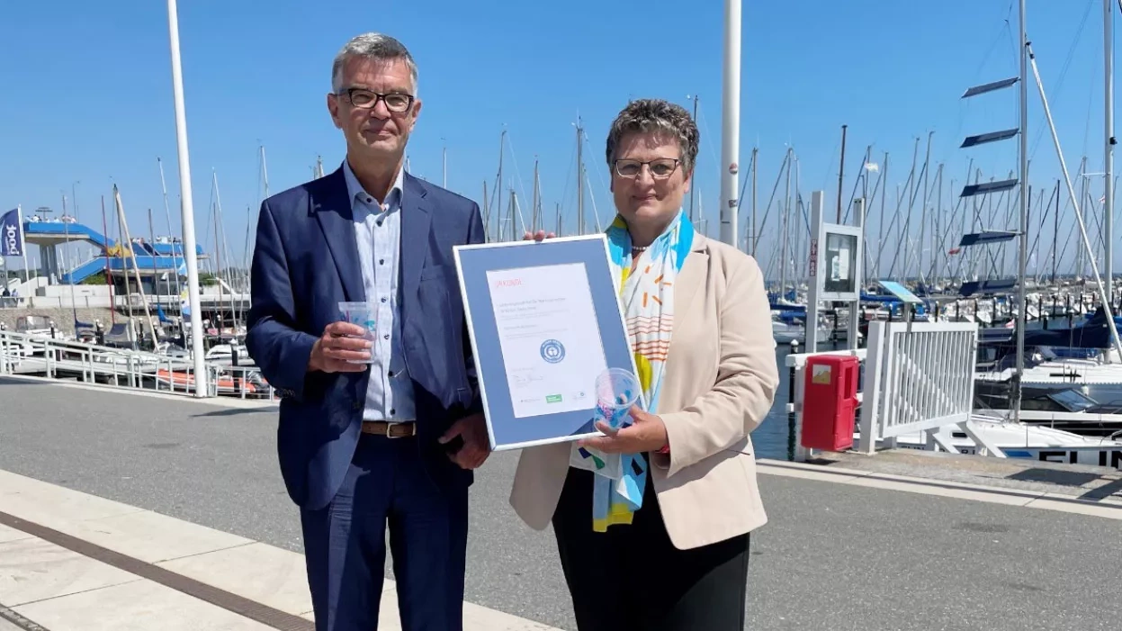 Mann mit Mehrwegbecher und eine Frau mit der Urkunde stehen an einem Hafen