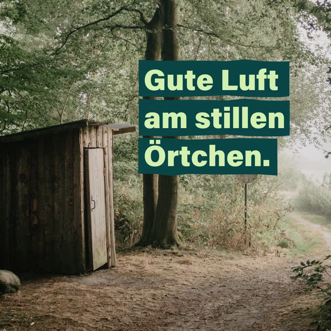 Ein hölzernes Toillettenhäuschen steht am Wegesrand unter einem Laubbaum. Auf dem Bild der Text Gute Luft am stillen Örtchen.