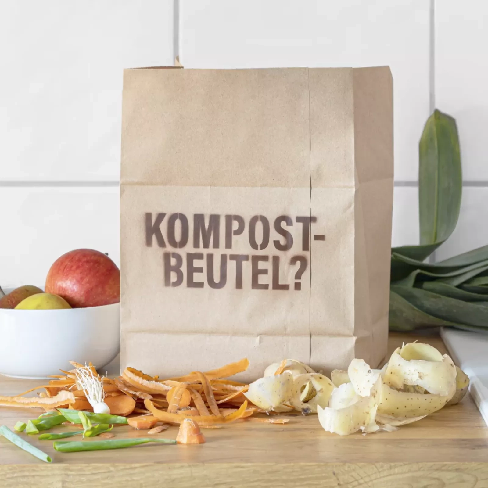 Ein Kompostbeutel aus Papier steht auf der Küchenablage. Das Wort "Kompostbeutel?" steht darauf geschrieben. Rund herum liegen Obst und verschiedene Gemüseschalen. 