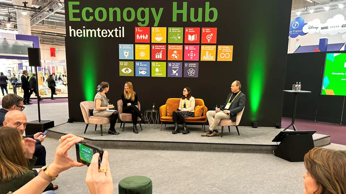 Drei Frauen und ein Mann im Gespräch auf einem Podium mit Publikum. Auf einer Stellwand steht Econogy Hub, heimtextil
