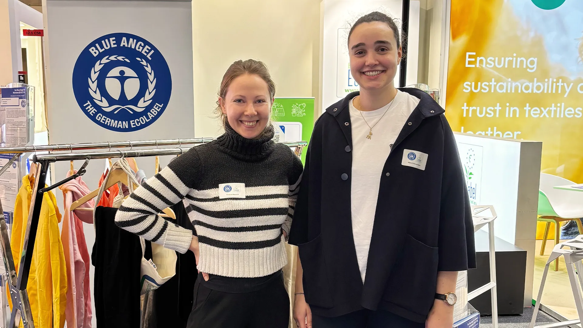 Zwei lächelnde Frauen stehen vor dem Blauen Engel Logo und einem Kleiderständer