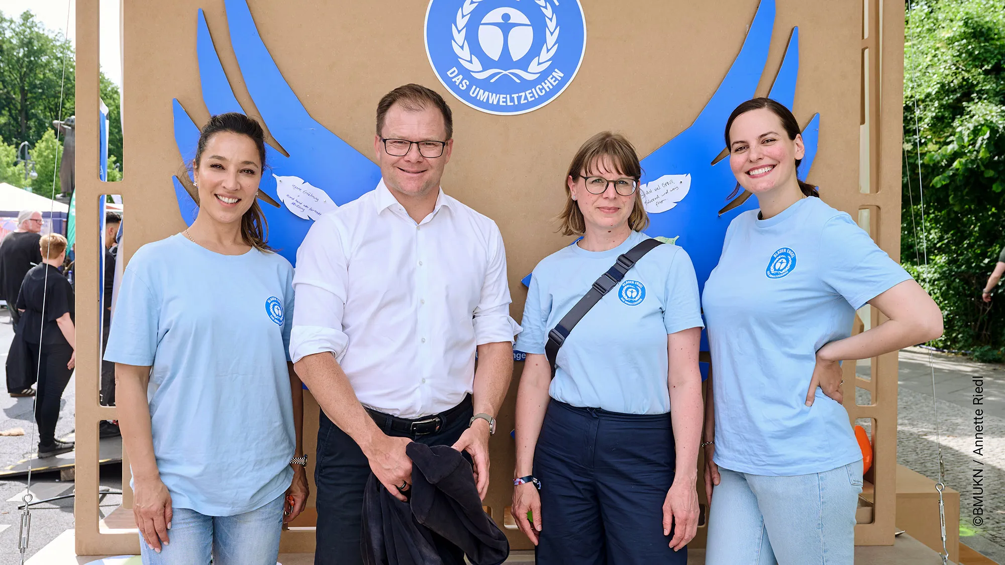 Foto von 4 Personen: Bundesumweltminister Carsten Schneider besucht den Blauer Engel-Stand, Im Hintergrund eine braune Wand mit Blauer Engel Logo und blauen Flügeln.