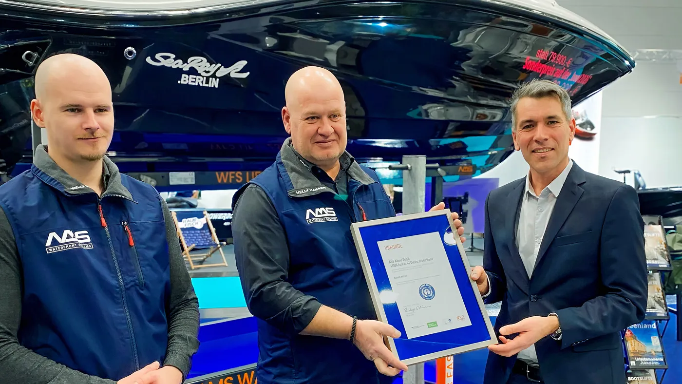 Zwei Männer in Westen von AMS und ein Mann im Anzug. Zwei halten gemeinsam die eingerahmte Zertifizierung. 