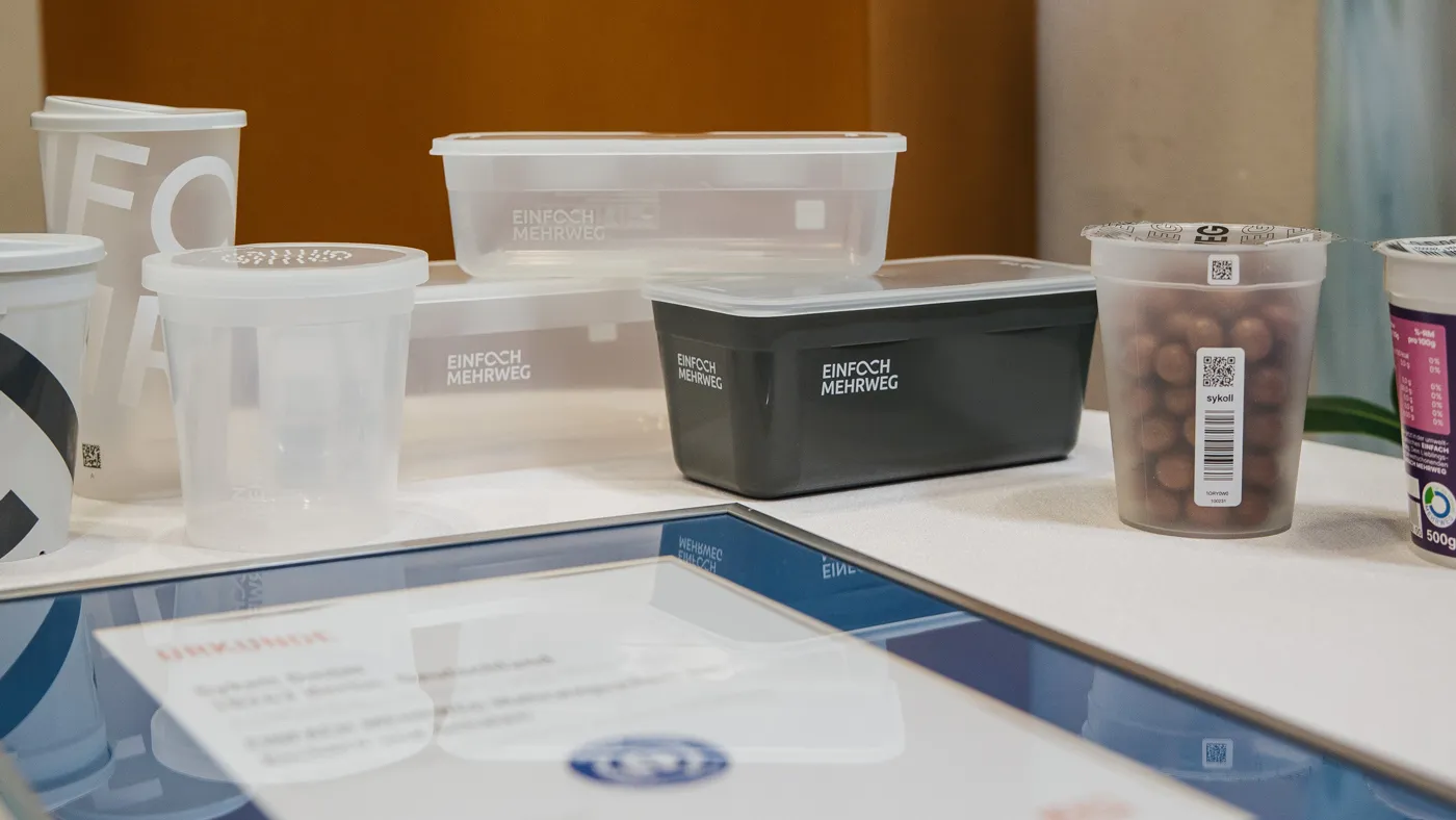 Ansicht von mehreren Mehrweg-Produktbehältern von Sykell neben der eingerahmten Urkunde vom Blauen Engel