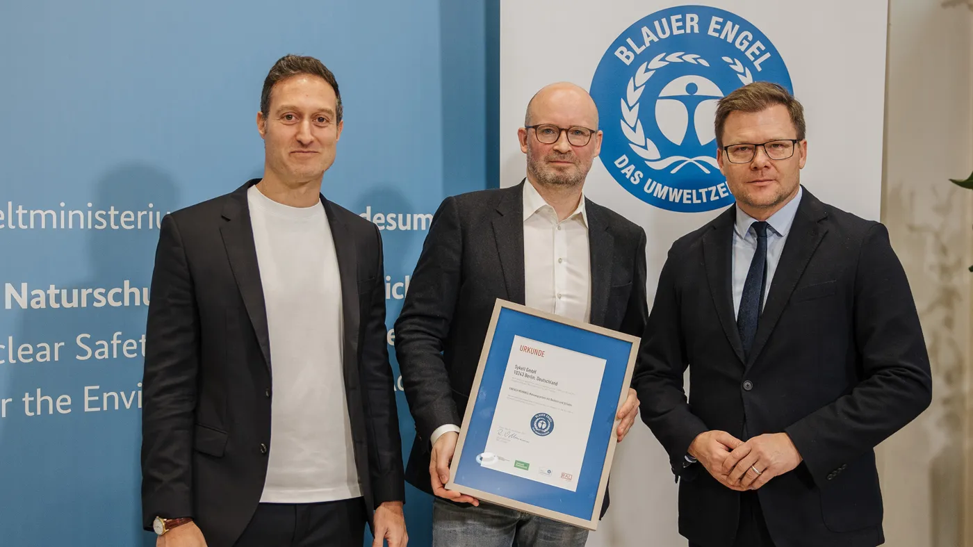 Zwei lächelnde Männer im Anzug, der eine hält die eingerahmte Zertifizierung durch den Blauen Engel in den Händen. Daneben steht freundlich blickend Bundesumweltminister Carsten Schneider.