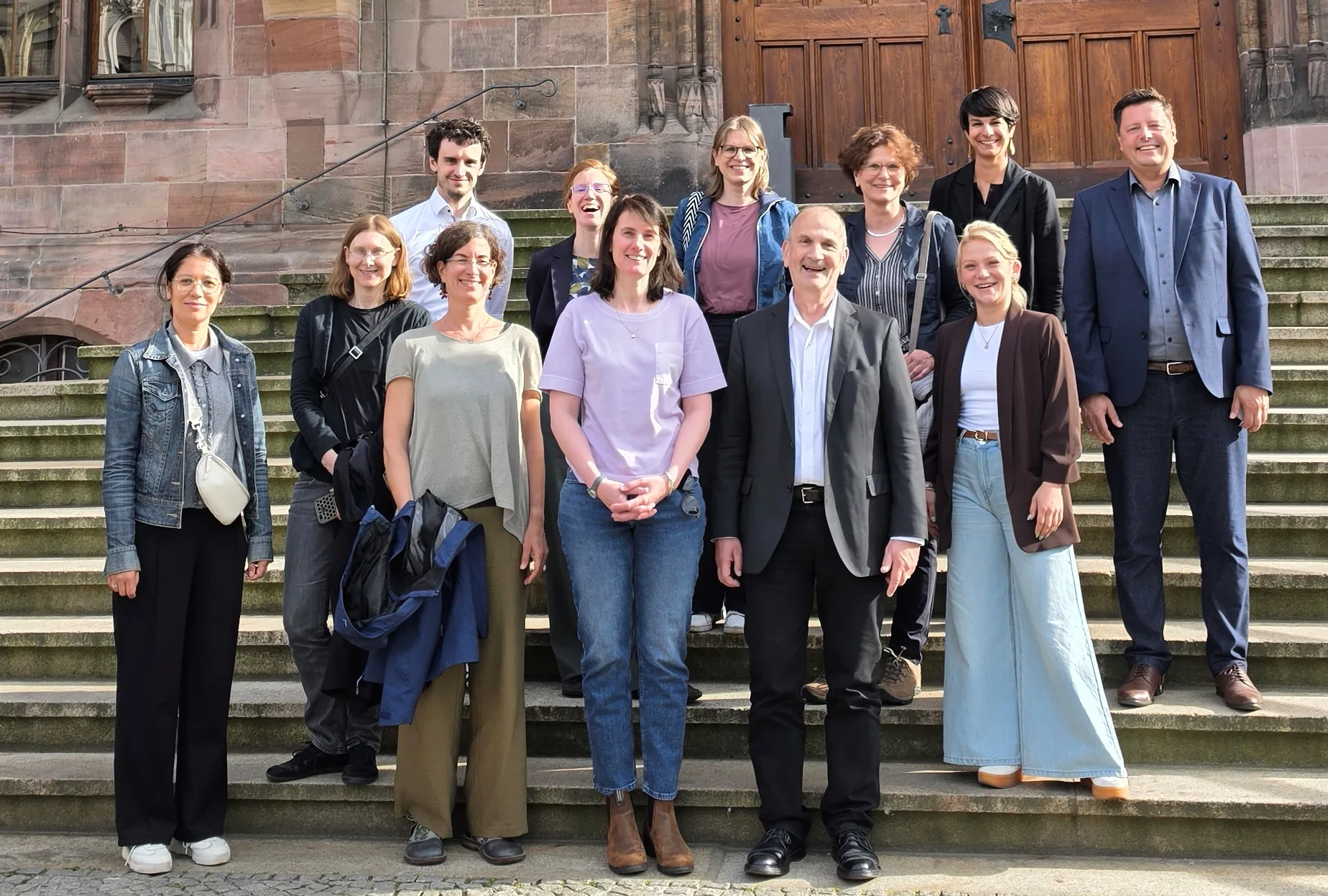 Eine Gruppe von fröhlich wirkenden Menschen steht auf den Stufen einer Sandsteinkirche