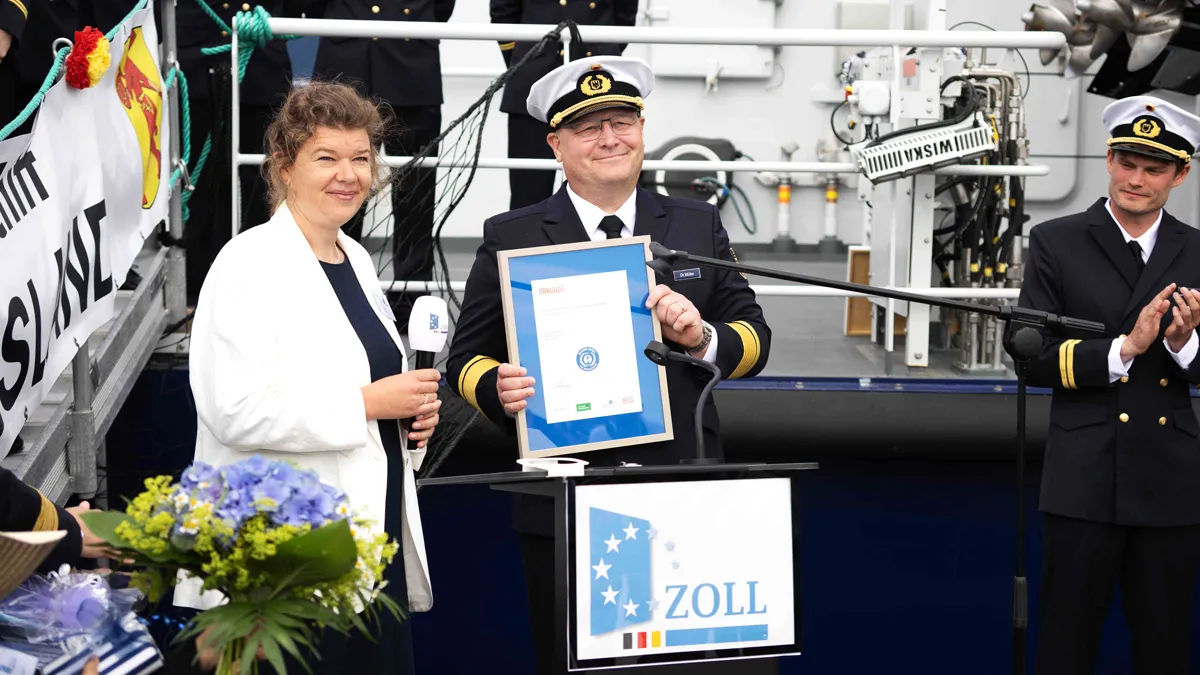 Frau Buttner und Herr Müller stehen hinter einem Pult mit der Aufschrift Zoll. Herr Müller mit Uniform hält die eingerahmte Zertifizierung hoch, daneben steht Herr Unnold auch in Uniform.  