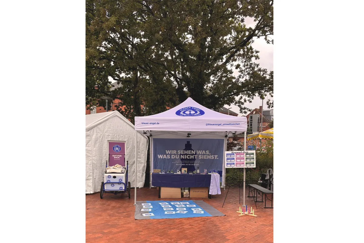 Stand des Blauen Engels unter Bäumen. Im hinteren Bereich ein Banner mit den Text "Wir sehen was, was du nicht siehst."