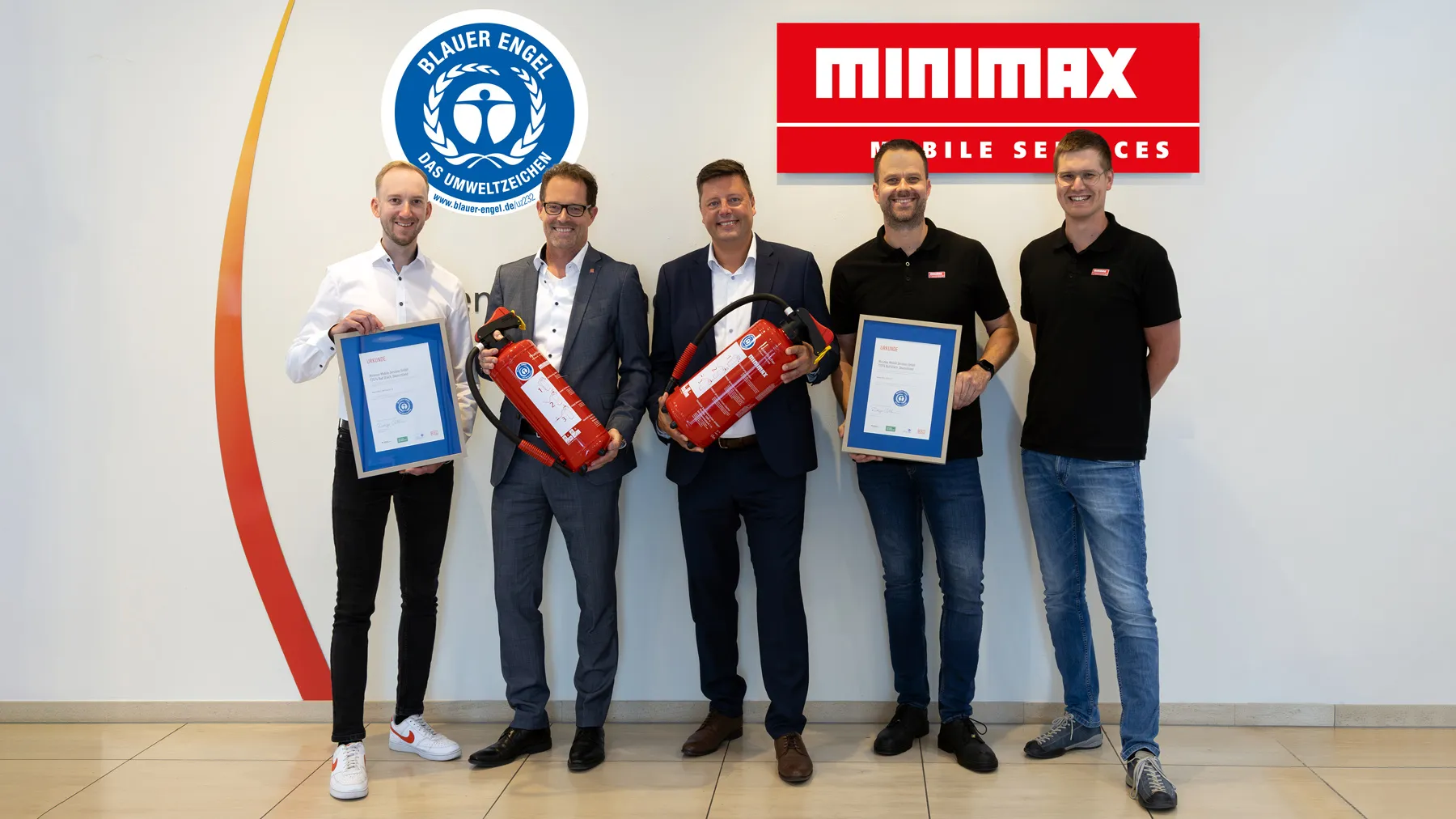 Fünf Männder stehen lächelnd mit zwei eingerahmten Urkunden und zwei Feuerlöschern vor einer Wand mit einem Logo des Blauen Engels und dem Logo Minimax - Mobile Services. 